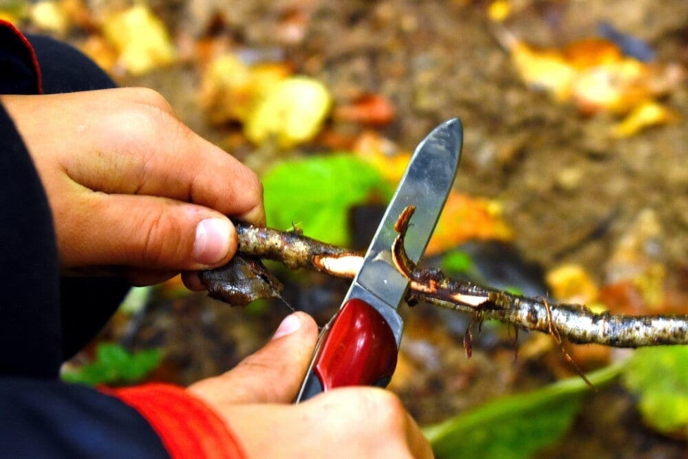 A boy with a knife cleans a stick in the forest. One child plays with a penknife.